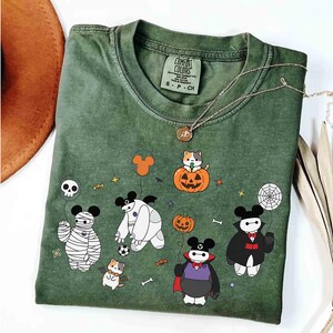 May include: Green Comfort Colors t-shirt featuring Halloween-themed cartoon characters. The design includes a mummy, a vampire, and a cat in a pumpkin. A gold necklace and a brown hat are also visible.