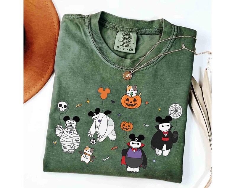 May include: Green Comfort Colors t-shirt featuring Halloween-themed cartoon characters. The design includes a mummy, a vampire, and a cat in a pumpkin. A gold necklace and a brown hat are also visible.