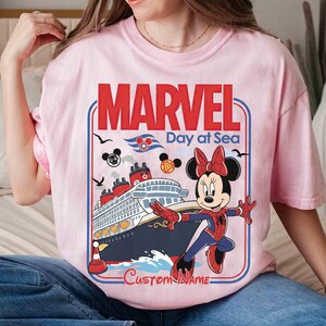 May include: A light pink t-shirt featuring a graphic of Minnie Mouse in a Spider-Man costume in front of a cruise ship. The shirt has the words "MARVEL Day at Sea" in red and blue, with a space for a custom name.