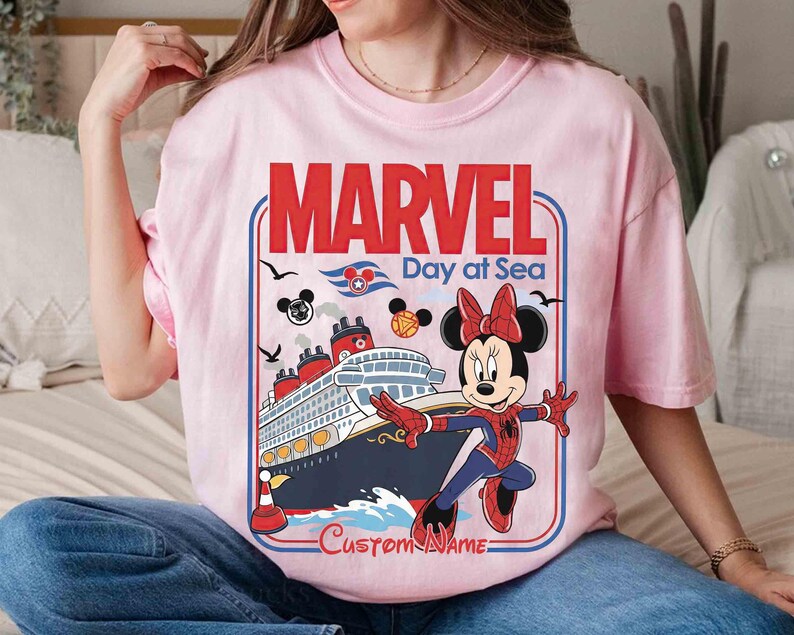 May include: A light pink t-shirt featuring a graphic of Minnie Mouse in a Spider-Man costume in front of a cruise ship. The shirt has the words "MARVEL Day at Sea" in red and blue, with a space for a custom name.