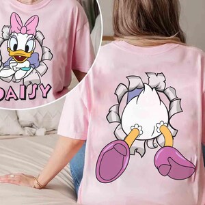 Puede incluir: Camiseta rosa claro con un dise&ntilde;o de Daisy Duck. La parte delantera de la camiseta muestra a Daisy rompiendo un efecto de papel rasgado, con su nombre debajo. La parte trasera de la camiseta muestra el trasero y los pies de Daisy saliendo del mismo efecto.