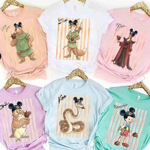 May include: A collection of six short-sleeve t-shirts in pastel colors, each featuring a Disney character wearing Mickey Mouse ears and holding a chocolate treat. Characters include Little John, Robin Hood, Jafar, Owl, Kaa, and Pinocchio.