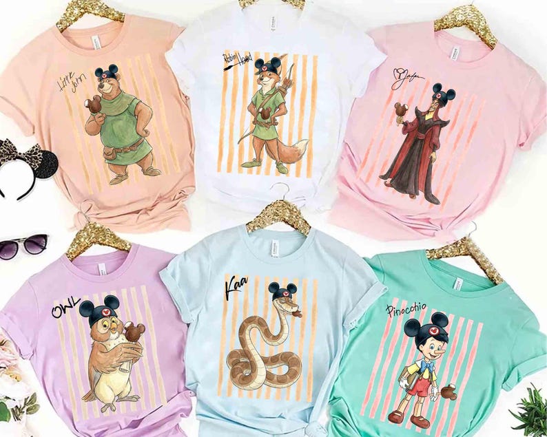 May include: A collection of six short-sleeve t-shirts in pastel colors, each featuring a Disney character wearing Mickey Mouse ears and holding a chocolate treat. Characters include Little John, Robin Hood, Jafar, Owl, Kaa, and Pinocchio.