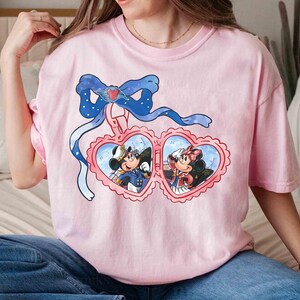 May include: Pink t-shirt with a graphic of Mickey Mouse and Minnie Mouse in a heart-shaped locket. The locket is hanging from a blue ribbon with white polka dots.