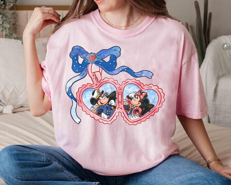 May include: Pink t-shirt with a graphic of Mickey Mouse and Minnie Mouse in a heart-shaped locket. The locket is hanging from a blue ribbon with white polka dots.