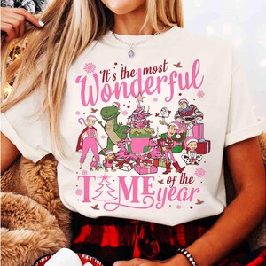 Puede incluir: Camiseta color crema con un gr&aacute;fico rosa que dice "It's the most Wonderful TIME of the year". El gr&aacute;fico presenta personajes de dibujos animados, un &aacute;rbol de Navidad y regalos. Un dise&ntilde;o festivo navide&ntilde;o.