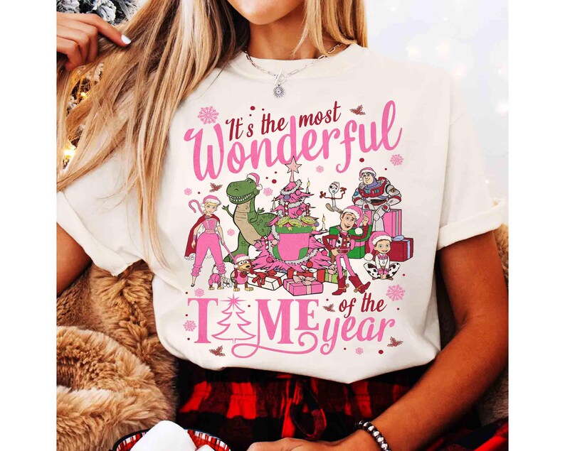 Puede incluir: Camiseta color crema con un gr&aacute;fico rosa que dice "It's the most Wonderful TIME of the year". El gr&aacute;fico presenta personajes de dibujos animados, un &aacute;rbol de Navidad y regalos. Un dise&ntilde;o festivo navide&ntilde;o.