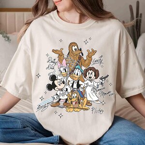 May include: Cream-colored t-shirt featuring a cartoon illustration of Disney characters in Star Wars-themed costumes. Mickey Mouse holds a lightsaber, while Minnie Mouse is dressed as Princess Leia. The shirt includes character names and star graphics.
