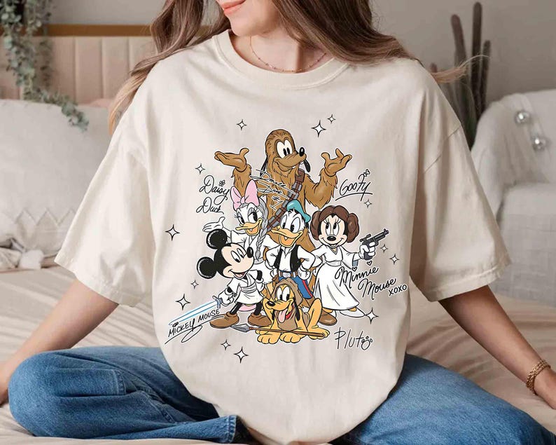 May include: Cream-colored t-shirt featuring a cartoon illustration of Disney characters in Star Wars-themed costumes. Mickey Mouse holds a lightsaber, while Minnie Mouse is dressed as Princess Leia. The shirt includes character names and star graphics.