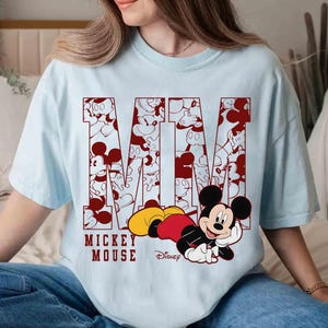 May include: Light blue t-shirt featuring the word "Mickey Mouse" in maroon, with the letters filled with Mickey Mouse illustrations. A cartoon Mickey Mouse is lying down on the bottom of the shirt.