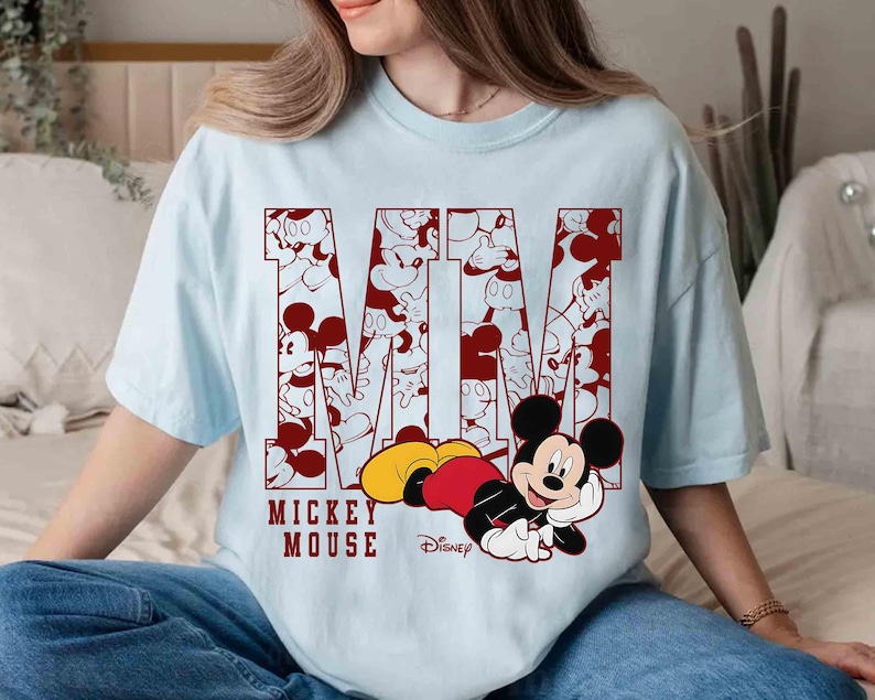 May include: Light blue t-shirt featuring the word "Mickey Mouse" in maroon, with the letters filled with Mickey Mouse illustrations. A cartoon Mickey Mouse is lying down on the bottom of the shirt.