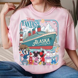 May include: Pink t-shirt featuring a Disney Fantasy Alaska cruise design. The graphic includes a cruise ship, cartoon characters, mountains, and the text "Disney Fantasy Alaska".