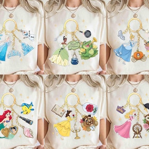 May include: Cream-colored t-shirts featuring various Disney princess-themed keychains. Each keychain showcases a different princess with related icons, such as a mermaid, a glass slipper, and a rose. The designs are colorful and whimsical.