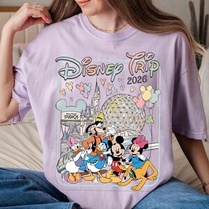 May include: A light purple t-shirt featuring a colorful graphic of Disney characters, including Mickey and Minnie Mouse, Goofy, Donald Duck, and Pluto. The graphic also includes the text "Disney Trip 2026" and various Disney park landmarks.