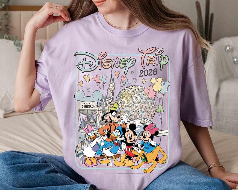 May include: A light purple t-shirt featuring a colorful graphic of Disney characters, including Mickey and Minnie Mouse, Goofy, Donald Duck, and Pluto. The graphic also includes the text "Disney Trip 2026" and various Disney park landmarks.