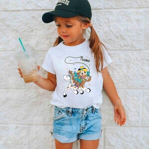 K&ouml;nnte beinhalten: Ein wei&szlig;es T-Shirt mit einem Cartoon-Cowboy auf einem Pferd und dem Wort "Name" dar&uuml;ber. Das Outfit umfasst Jeansshorts und eine schwarze Baseballkappe. Das M&auml;dchen h&auml;lt einen durchsichtigen Plastikbecher mit einem Strohhalm.