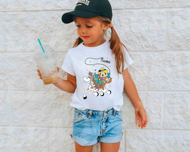 K&ouml;nnte beinhalten: Ein wei&szlig;es T-Shirt mit einem Cartoon-Cowboy auf einem Pferd und dem Wort "Name" dar&uuml;ber. Das Outfit umfasst Jeansshorts und eine schwarze Baseballkappe. Das M&auml;dchen h&auml;lt einen durchsichtigen Plastikbecher mit einem Strohhalm.