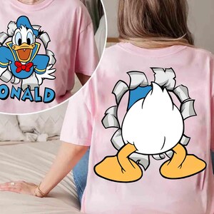 May include: Pink t-shirt featuring a graphic of Donald Duck bursting through a torn background. The front of the shirt displays Donald with the name "DONALD" in blue. The back shows Donald's rear and feet.