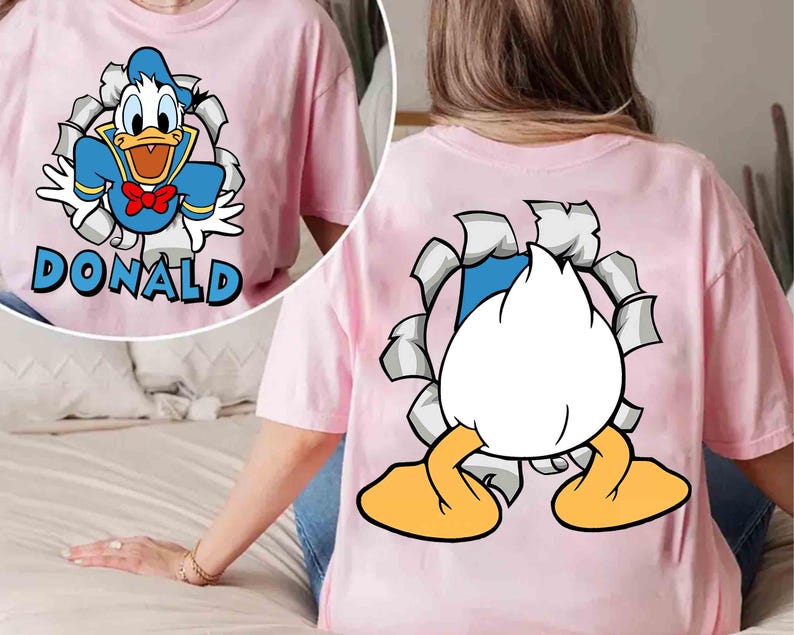 May include: Pink t-shirt featuring a graphic of Donald Duck bursting through a torn background. The front of the shirt displays Donald with the name "DONALD" in blue. The back shows Donald's rear and feet.