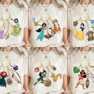 May include: Cream-colored t-shirts featuring various Disney character keychains. Each keychain includes a character and related items, such as a magic lamp, a tiger, a well, a bow and arrow, a dragon, and a pig. The designs are colorful and detailed.