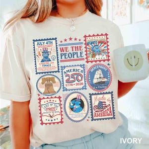 以下が含まれることがあります： アイボリー色のTシャツ。愛国的な切手のデザインのコラージュが特徴です。切手には、自由の鐘、船、そして「7月4日」と「アメリカ」の文字が描かれています。Tシャツには「We the People」の文字も入っています。