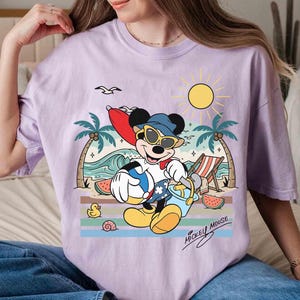 K&ouml;nnte beinhalten: Ein helllila T-Shirt mit einem Cartoon von Mickey Mouse am Strand. Mickey tr&auml;gt eine Sonnenbrille und einen Hut und h&auml;lt einen Strandball und einen Eimer. Das Design umfasst Palmen, eine Sonne, Wellen und den Text "Mickey Mouse".