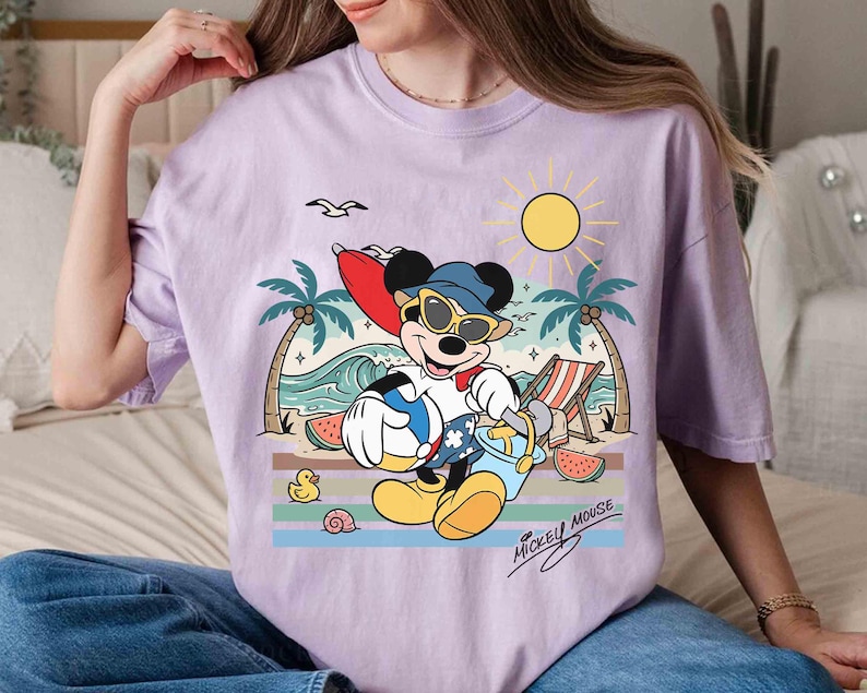 K&ouml;nnte beinhalten: Ein helllila T-Shirt mit einem Cartoon von Mickey Mouse am Strand. Mickey tr&auml;gt eine Sonnenbrille und einen Hut und h&auml;lt einen Strandball und einen Eimer. Das Design umfasst Palmen, eine Sonne, Wellen und den Text "Mickey Mouse".