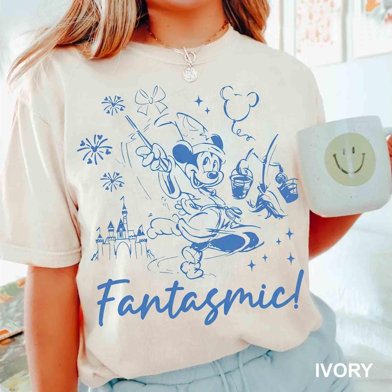Puede incluir: Camiseta de color marfil con un gr&aacute;fico azul de Mickey Mouse como hechicero, con la palabra "Fantasmic!" debajo. El dise&ntilde;o incluye fuegos artificiales, un castillo y una taza con una carita sonriente. La camiseta es de material suave.