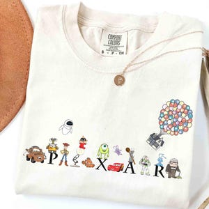 K&ouml;nnte beinhalten: Cremefarbenes T-Shirt mit dem Wort "PIXAR" und Zeichentrickfiguren aus verschiedenen Pixar-Filmen. Eine goldene Halskette und ein brauner Hut sind ebenfalls zu sehen. Das Shirt besteht aus einem weichen Material.