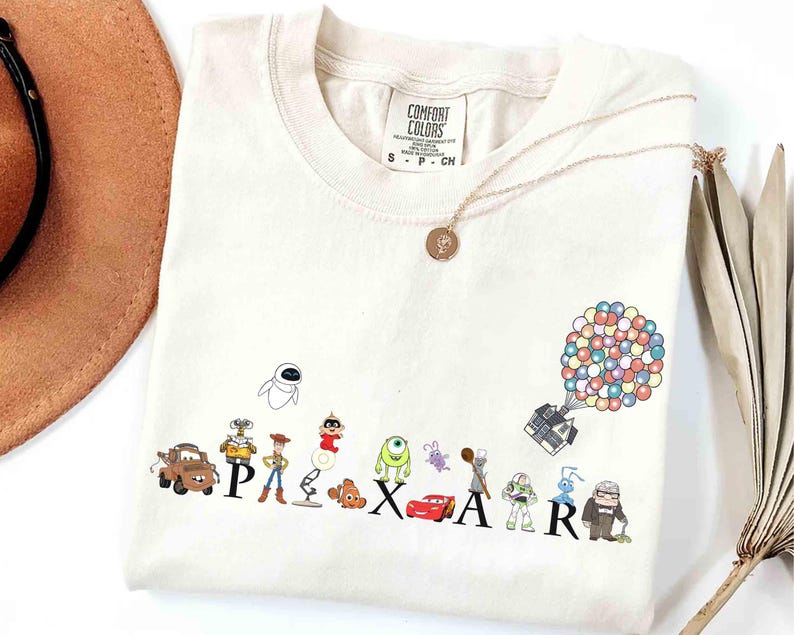 K&ouml;nnte beinhalten: Cremefarbenes T-Shirt mit dem Wort "PIXAR" und Zeichentrickfiguren aus verschiedenen Pixar-Filmen. Eine goldene Halskette und ein brauner Hut sind ebenfalls zu sehen. Das Shirt besteht aus einem weichen Material.