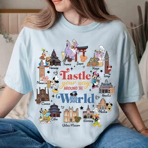 May include: Light blue t-shirt featuring a colorful graphic with the text "Tastle your way around the World." The design includes cartoon characters and landmarks from various countries, such as France, Japan, and Italy.