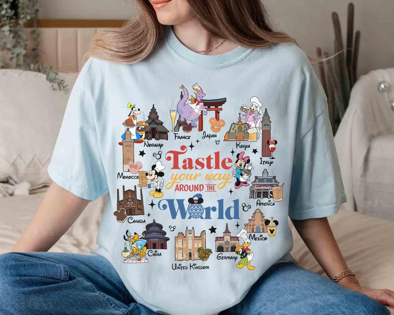 May include: Light blue t-shirt featuring a colorful graphic with the text "Tastle your way around the World." The design includes cartoon characters and landmarks from various countries, such as France, Japan, and Italy.