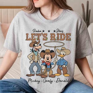 May include: A gray t-shirt featuring Mickey Mouse, Goofy, and Donald Duck dressed as cowboys with the text "Rodeo Stars Let's Ride". The characters are wearing cowboy hats, vests, and holding ropes.