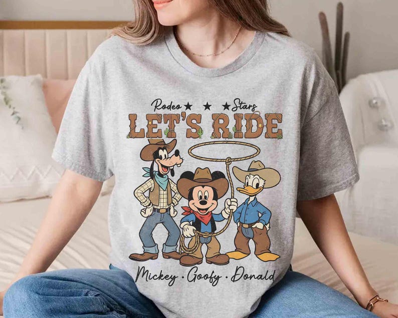 May include: A gray t-shirt featuring Mickey Mouse, Goofy, and Donald Duck dressed as cowboys with the text "Rodeo Stars Let's Ride". The characters are wearing cowboy hats, vests, and holding ropes.
