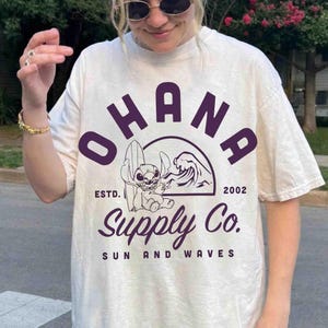 May include: Cream-colored t-shirt featuring the word "OHANA" in purple block letters above a graphic of a cartoon character with a surfboard and a wave. The text "Supply Co. SUN AND WAVES" is below the graphic. The shirt has short sleeves.