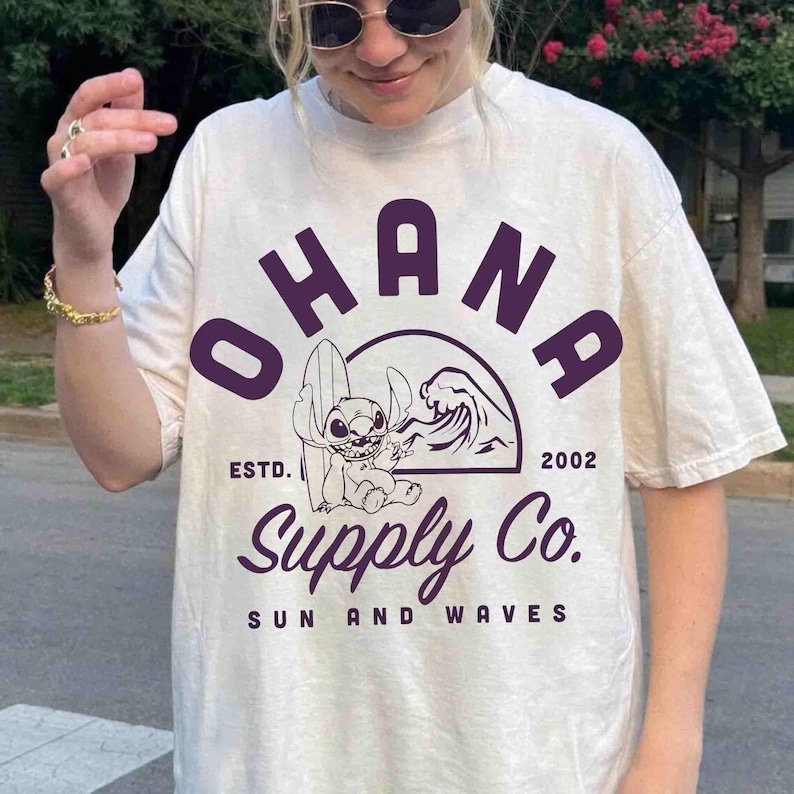 May include: Cream-colored t-shirt featuring the word "OHANA" in purple block letters above a graphic of a cartoon character with a surfboard and a wave. The text "Supply Co. SUN AND WAVES" is below the graphic. The shirt has short sleeves.