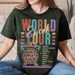 K&ouml;nnte beinhalten: Ein gr&uuml;nes T-Shirt mit einer bunten Grafik, auf der "World Epcot Tour" steht und L&auml;nder wie Kanada, Vereinigtes K&ouml;nigreich, Frankreich, Marokko, China, Norwegen, Mexiko, Japan, Vereinigte Staaten, Italien und Deutschland aufgelistet sind.