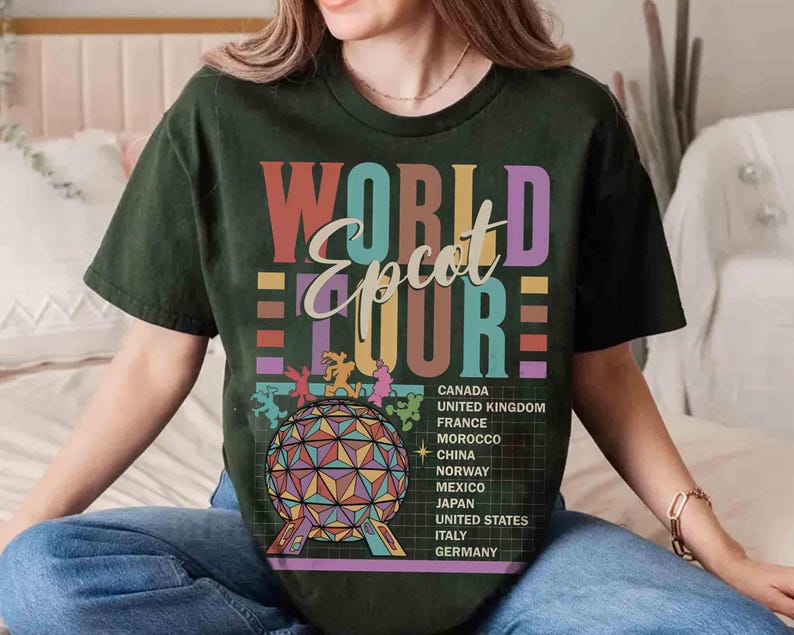 K&ouml;nnte beinhalten: Ein gr&uuml;nes T-Shirt mit einer bunten Grafik, auf der "World Epcot Tour" steht und L&auml;nder wie Kanada, Vereinigtes K&ouml;nigreich, Frankreich, Marokko, China, Norwegen, Mexiko, Japan, Vereinigte Staaten, Italien und Deutschland aufgelistet sind.
