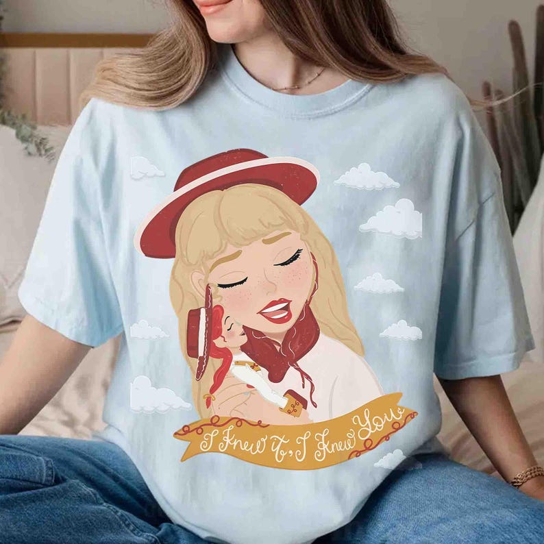 May include: A light blue t-shirt featuring a graphic illustration of a blonde character wearing a red hat and holding a smaller doll. The design includes white clouds and a yellow banner at the bottom with the text I Knew It, I Knew You.