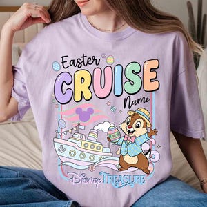 K&ouml;nnte beinhalten: Helllila T-Shirt mit der Aufschrift "Easter CRUISE Name" in Pastellfarben. Eine Zeichentrickfigur h&auml;lt ein Osterei in der N&auml;he eines Schiffs, darunter steht "Disney TREASURE".