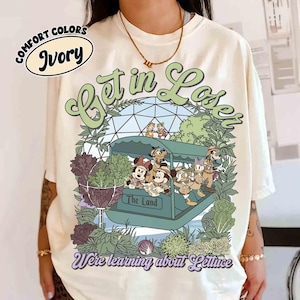 May include: Ivory-colored t-shirt with a graphic featuring Disney characters in a boat labeled "The Land." The design includes the text "Get in Lose" and "We're learning about Lettuce" in green and purple. The shirt is from Comfort Colors.