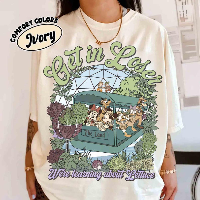 May include: Ivory-colored t-shirt with a graphic featuring Disney characters in a boat labeled "The Land." The design includes the text "Get in Lose" and "We're learning about Lettuce" in green and purple. The shirt is from Comfort Colors.