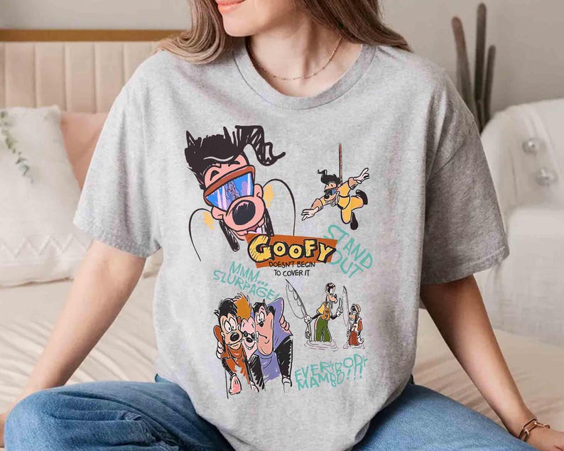 May include: Grey t-shirt featuring a cartoon graphic of Goofy and other Disney characters. The text on the shirt reads "Goofy Doesn't Begin To Cover It" and "Stand Out".