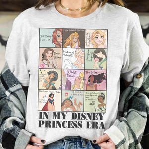 May include: A light gray t-shirt featuring a grid of Disney princess illustrations with quotes. The shirt has the text "IN MY DISNEY PRINCESS ERA" at the bottom. The shirt is worn by a person.