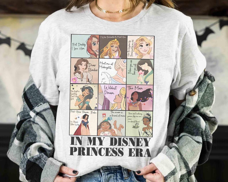 May include: A light gray t-shirt featuring a grid of Disney princess illustrations with quotes. The shirt has the text "IN MY DISNEY PRINCESS ERA" at the bottom. The shirt is worn by a person.