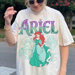 Op de afbeelding: Cr&egrave;mekleurig T-shirt met het woord "ARIEL" in paars en groen, met zeemeerminstaartaccenten. Het shirt toont een cartoonafbeelding van Ariel in een turquoise jurk, met rood haar en twee turquoise omtrektekens van Ariel.