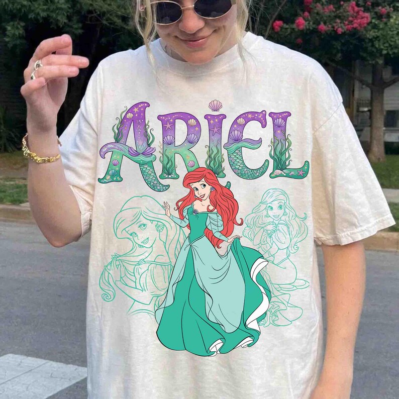 Op de afbeelding: Cr&egrave;mekleurig T-shirt met het woord "ARIEL" in paars en groen, met zeemeerminstaartaccenten. Het shirt toont een cartoonafbeelding van Ariel in een turquoise jurk, met rood haar en twee turquoise omtrektekens van Ariel.