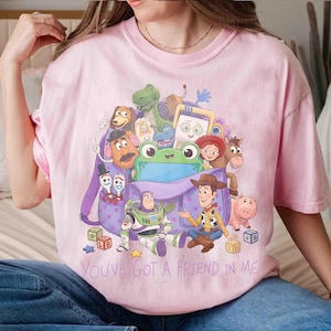 May include: A light pink t-shirt featuring a graphic print of various Toy Story characters emerging from a purple backpack. The characters include Woody, Buzz Lightyear, Rex, and others. The text "YOU'VE GOT A FRIEND IN ME" is printed below the graphic.