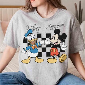 May include: A gray t-shirt featuring Donald Duck and Mickey Mouse. Donald Duck is wearing a blue sailor suit and Mickey Mouse is wearing red shorts. The text "Donald Duck #1" and "Mickey Mouse" are above the characters.