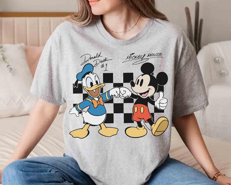 May include: A gray t-shirt featuring Donald Duck and Mickey Mouse. Donald Duck is wearing a blue sailor suit and Mickey Mouse is wearing red shorts. The text "Donald Duck #1" and "Mickey Mouse" are above the characters.